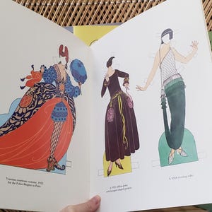Puede incluir: Libro abierto con ilustraciones de dise&ntilde;os de moda vintage. La p&aacute;gina izquierda presenta a una mujer con un vestido largo rojo con detalles azules y negros. La p&aacute;gina derecha muestra a dos mujeres con vestidos. Un libro verde titulado "Designs of Parties" es visible.