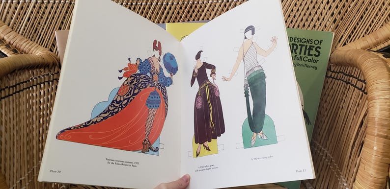 Puede incluir: Libro abierto con ilustraciones de dise&ntilde;os de moda vintage. La p&aacute;gina izquierda presenta a una mujer con un vestido largo rojo con detalles azules y negros. La p&aacute;gina derecha muestra a dos mujeres con vestidos. Un libro verde titulado "Designs of Parties" es visible.