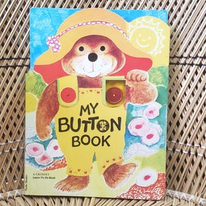 May include: A children's book titled "My Button Book" featuring a cartoon bear wearing a yellow hat and overalls. The cover has a button and a smiling sun illustration. The background is a woven pattern.