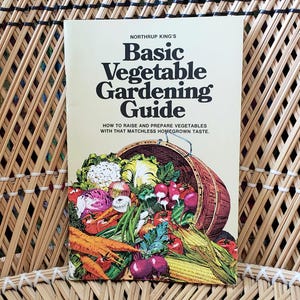 May include: A vintage gardening guide titled "Basic Vegetable Gardening Guide" by Northrup King's. The cover features a basket overflowing with colorful vegetables. The book is set against a woven wicker background.