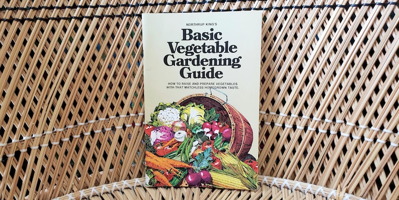 May include: A vintage gardening guide titled "Basic Vegetable Gardening Guide" by Northrup King's. The cover features a basket overflowing with colorful vegetables. The book is set against a woven wicker background.