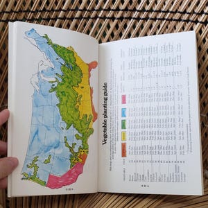 May include: Open book with a map of the United States, color-coded for vegetable planting zones. The left page shows a map with blue, green, yellow, and pink areas. The right page has a chart with text. The book is open on a woven surface.