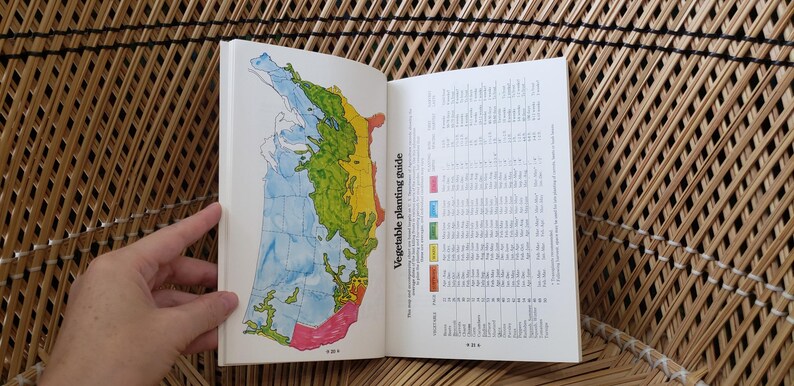 May include: Open book with a map of the United States, color-coded for vegetable planting zones. The left page shows a map with blue, green, yellow, and pink areas. The right page has a chart with text. The book is open on a woven surface.