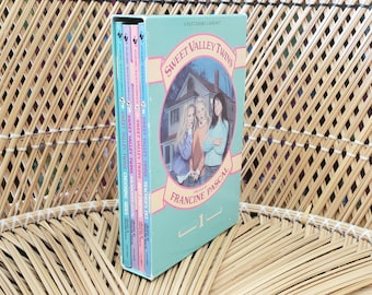 Puede incluir: Un set de libros Sweet Valley Twins de Francine Pascal, volumen 1. La caja azul claro presenta una ilustración de tres personas frente a una casa. El set contiene tres libros delgados con lomos azul claro, rosa y morado.