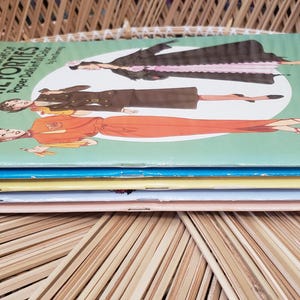 Puede incluir: Una pila de libros de moda vintage titulados "Great Fashion Designs of the Forties". Las portadas presentan ilustraciones de mujeres con atuendos de los a&ntilde;os 40. Los libros est&aacute;n apilados sobre una superficie de mimbre tejido.