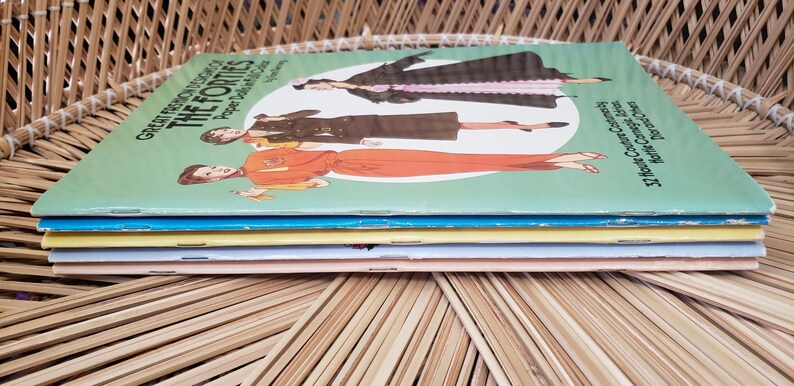 Puede incluir: Una pila de libros de moda vintage titulados "Great Fashion Designs of the Forties". Las portadas presentan ilustraciones de mujeres con atuendos de los a&ntilde;os 40. Los libros est&aacute;n apilados sobre una superficie de mimbre tejido.