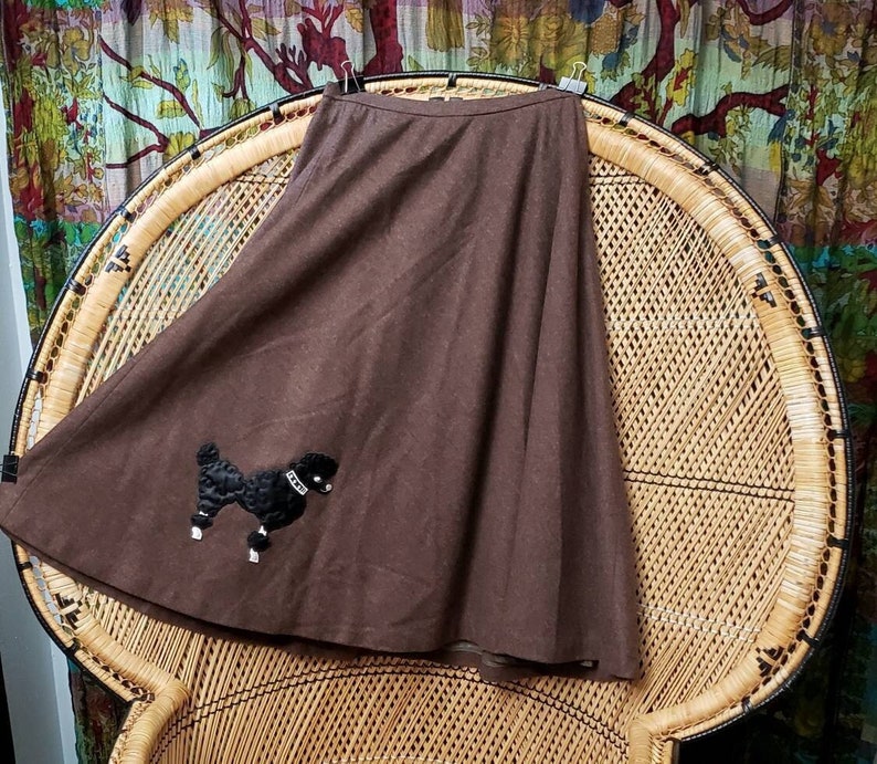 May include: A brown wool skirt with a black poodle applique. The skirt is a-line and has a high waist. It is sitting on a wicker chair.