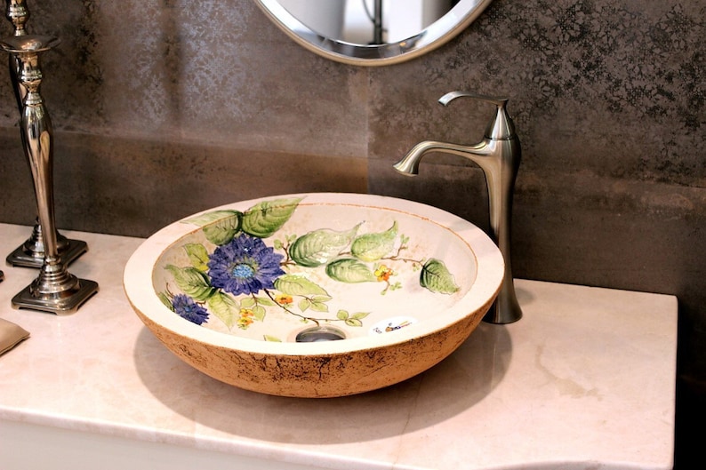 Puede incluir: Un lavabo de ba&ntilde;o de cer&aacute;mica blanca con un dise&ntilde;o floral con flores azules, verdes y amarillas. El lavabo tiene forma ovalada y una superficie rugosa y texturizada. Un grifo plateado est&aacute; conectado al lavabo.
