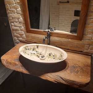 Peut inclure: Un lavabo de salle de bain en c&eacute;ramique blanche avec un robinet noir et un motif de branche d'olivier. Le lavabo est mont&eacute; sur un comptoir en bois rustique.