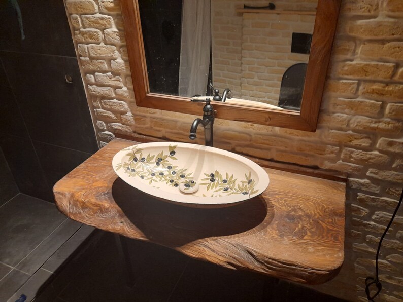 Peut inclure: Un lavabo de salle de bain en c&eacute;ramique blanche avec un robinet noir et un motif de branche d'olivier. Le lavabo est mont&eacute; sur un comptoir en bois rustique.