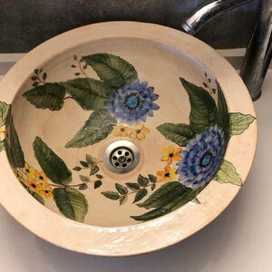 Puede incluir: Un lavabo de cer&aacute;mica blanca con un dise&ntilde;o floral. El lavabo presenta flores azules y amarillas con hojas verdes.
