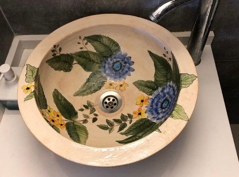 Puede incluir: Un lavabo de cer&aacute;mica blanca con un dise&ntilde;o floral. El lavabo presenta flores azules y amarillas con hojas verdes.