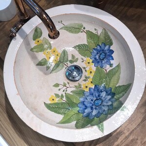 Puede incluir: Un lavabo de ba&ntilde;o de cer&aacute;mica blanca con un dise&ntilde;o floral. El lavabo presenta flores azules y amarillas con hojas verdes. El lavabo tiene un borde blanco y un desag&uuml;e cromado.