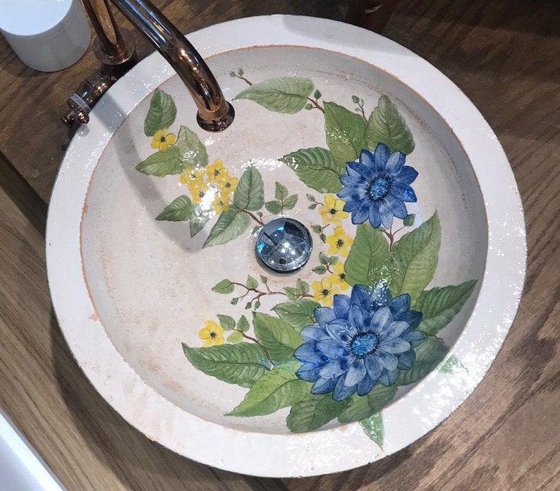 Puede incluir: Un lavabo de ba&ntilde;o de cer&aacute;mica blanca con un dise&ntilde;o floral. El lavabo presenta flores azules y amarillas con hojas verdes. El lavabo tiene un borde blanco y un desag&uuml;e cromado.