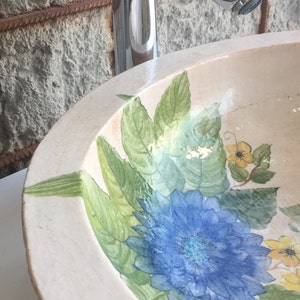 Puede incluir: Un lavabo de cer&aacute;mica blanca con un dise&ntilde;o floral. El lavabo presenta flores y hojas azules, verdes y amarillas.