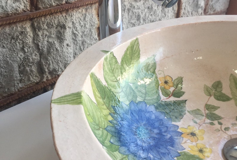 Puede incluir: Un lavabo de cer&aacute;mica blanca con un dise&ntilde;o floral. El lavabo presenta flores y hojas azules, verdes y amarillas.