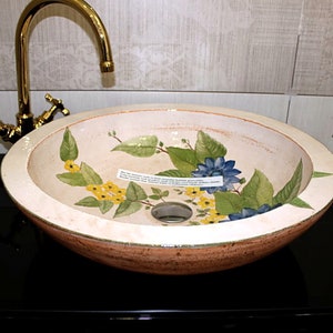 Puede incluir: Un lavabo de ba&ntilde;o de cer&aacute;mica blanca con un dise&ntilde;o floral con flores amarillas, azules y verdes. El lavabo es redondo y tiene un desag&uuml;e en el centro.