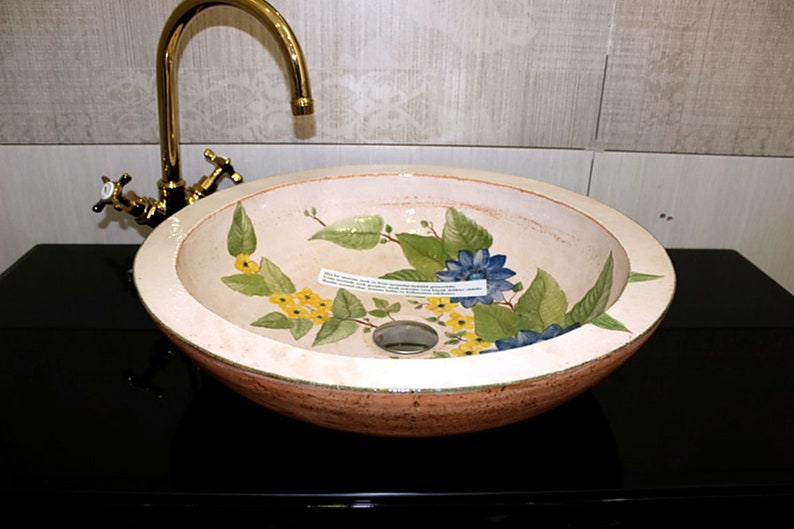 Puede incluir: Un lavabo de ba&ntilde;o de cer&aacute;mica blanca con un dise&ntilde;o floral con flores amarillas, azules y verdes. El lavabo es redondo y tiene un desag&uuml;e en el centro.