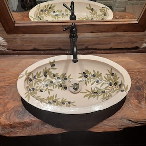 Peut inclure: Un lavabo en c&eacute;ramique ovale blanc avec un robinet noir et un motif de branche d'olivier. Le lavabo est mont&eacute; sur un comptoir en bois.