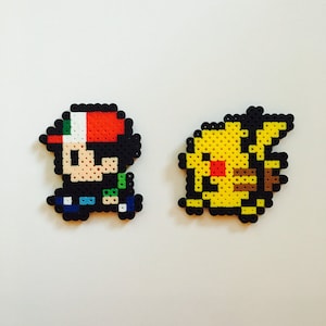 May include: Two pixel art figures: a character with a red, white, and green hat and a yellow Pikachu. The figures are made of small, colorful beads, likely for crafting or decoration. The background is plain white.