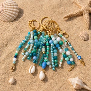 May include: A collection of beaded keychains with gold-colored hardware. The keychains feature various shades of blue, green, and white beads, with seashell and charm accents. The items are arranged on a sandy surface, with seashells and a starfish.