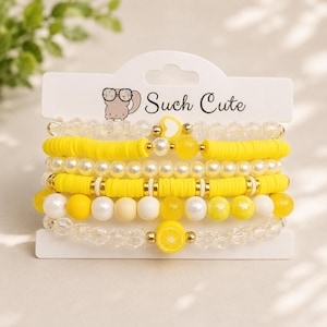 May include: A set of five stacked bracelets on a white display card with the text Such Cute. The bracelets feature yellow clay discs, white faux pearls, clear faceted beads, and yellow beads, including a heart charm and a lemon slice charm.