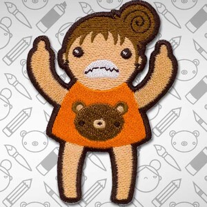 May include: Embroidered iron-on patch of a cartoon girl with brown hair, wearing an orange shirt with a brown bear on it, and raising her arms in the air. The girl has a grumpy expression on her face.