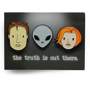 May include: Enamel pins featuring cartoon depictions of two people and an alien, with the text "the truth is out there." The pins have a black background. The person on the left has brown hair, the alien is gray, and the person on the right has orange hair.
