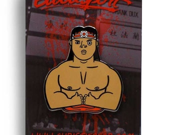 May include: A red, white, and black enamel pin featuring a cartoon character with a red headband and a red circle on his forehead. The character is wearing a red shirt and has a muscular build. The pin is set against a black background with red and white text that reads "Bloodsport".