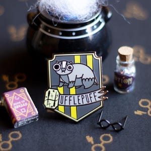 May include: A gold and yellow enamel pin featuring the Hufflepuff house crest from the Harry Potter series. The crest depicts a badger with a book and the words "Hufflepuff" on a banner.
