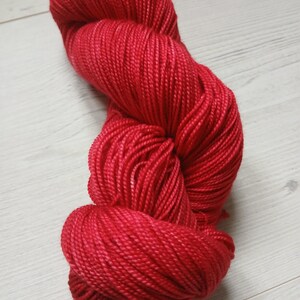 May include: A bright red skein of yarn, tightly twisted and textured. The yarn has a solid colour with slight tonal variations. The image is a close-up, highlighting the fine strands and the overall shape. Suitable for knitting or crochet projects, this yarn is a rich crimson.