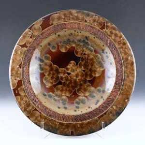 May include: A brown ceramic plate with a decorative design of brown and gold circles and swirls. The plate has a raised rim and a glossy finish.