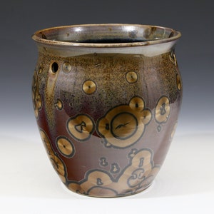 Puede incluir: Una maceta de cerámica marrón con un esmalte moteado y detalles dorados. La maceta tiene un cuerpo redondo y una abertura ancha.