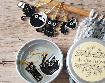 May include: A set of four black cat knitting stitch markers in a silver tin with a label that says "Knitting Notions". The markers are in the shape of a cat, a cat in a bowl, a cat with a ball of yarn, and a cat with a hat. The tin is sitting on a wooden surface next to a piece of grey knitted fabric.