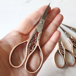 May include: A set of five ornate scissors with copper-toned handles and silver blades. The scissors have intricate designs on the handles and are held in a hand.
