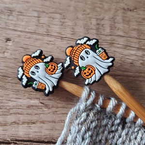May include: Two wooden knitting needles with Halloween-themed ghost and pumpkin charms. The charms are orange, white, and black. The needles are resting on a gray knitted fabric.