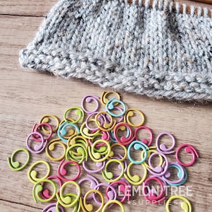 May include: A close-up of a pile of colourful plastic stitch markers in various shades of yellow, green, pink, and purple. The markers are in the shape of a circle with a small tail. The text "LEMON TREE SUPPLIES" is visible in the bottom right corner of the image.