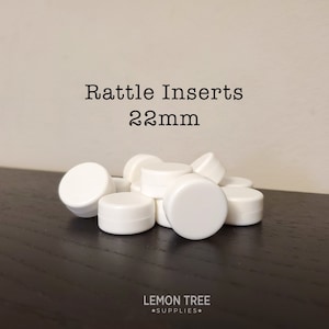 May include: A pile of white plastic rattle inserts, 22mm in diameter.  Rattle Inserts 22mm is written in black text above the pile.  The inserts are on a dark wood surface.