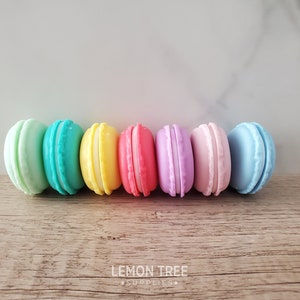 May include: A row of six pastel-coloured macarons, in shades of green, blue, yellow, pink, purple, and light blue, arranged in a line on a wooden surface.
