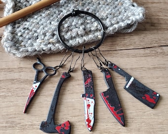 May include: A set of six black metal stitch markers shaped like knives and an axe, with red and white paint to look like blood splatters. The markers are attached to a black metal ring. The markers are on a wooden surface with a piece of grey knitted fabric and a wooden knitting needle in the background. The text "LEMON TREE SUPPLIES" is visible at the bottom of the image.