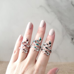 May include: Three silver snake rings with colourful enamel details. The rings are on a hand, with the green snake ring on the middle finger, the red snake ring on the index finger, and the black snake ring on the ring finger.