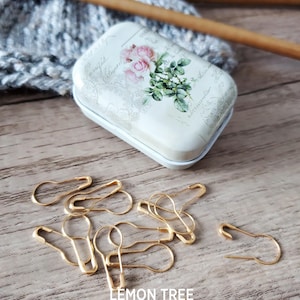 May include: A white metal tin with a floral design and the text "Lemon Tree Supplies" on a wooden surface. The tin is open and contains a group of gold safety pins.