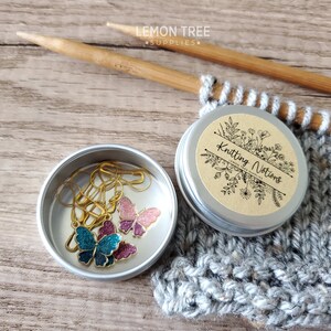 May include: A small silver tin with a brown label that says "Knitting Notions" with a floral design. The tin contains three gold butterfly stitch markers, one blue, one pink, and one purple.