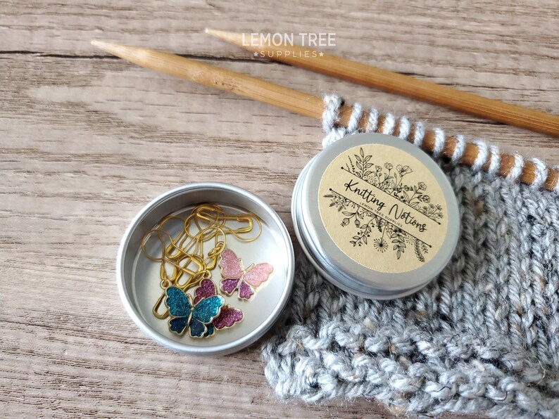 May include: A small silver tin with a brown label that says "Knitting Notions" with a floral design. The tin contains three gold butterfly stitch markers, one blue, one pink, and one purple.