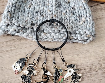 May include: Five black and gold stitch markers with different designs, including a skull and moon, a crow, a plague doctor, and a crow with a scythe. The markers are attached to a black ring.