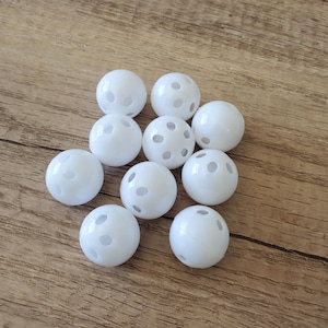 May include: Ten white plastic balls with evenly spaced holes are displayed on a wooden surface. The balls are round and smooth, suggesting their use in sports or games. The holes are a key feature of the design.