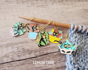 May include: A selection of enamel cat stitch markers in various designs. The markers depict black cats in pots, baskets, and a cactus, with gold-coloured hooks. The image includes a wooden knitting needle and a knitted swatch.