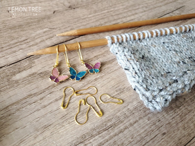 May include: Three butterfly-shaped stitch markers in pink, blue, and purple with gold accents. The markers are hanging from a wooden knitting needle. There are also five gold safety pins on a wooden surface.