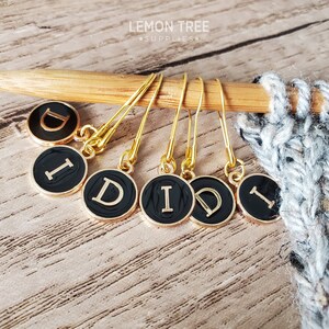 May include: Six gold stitch markers with black round charms featuring a gold letter "D" in a typewriter font. The markers are hanging on a wooden knitting needle.
