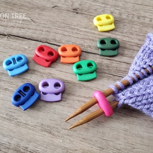 May include: Assortment of colorful plastic toggles, also known as cord locks, in various colors including blue, green, purple, orange, red, and yellow. The toggles are used for securing cords and ropes.  A purple knitted fabric and wooden knitting needles are also in the image.